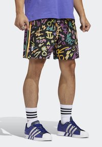 Pantaloncini colorati con motivi astratti in rosa, giallo e blu su uno sfondo nero, abbinati a scarpe blu e calzini bianchi a righe.