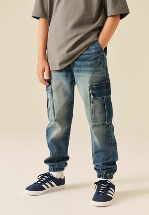 Child wearing faded blue denim cargo jogger pants, grey t-shirt, white socks, and navy blue sneakers with white stripes, standing with hands in pockets.