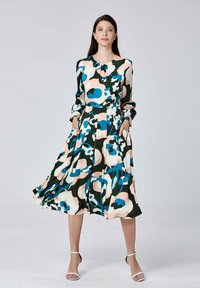 Femme portant une robe à manches longues, arrivant aux genoux, avec des motifs abstraits bleus, roses, noirs et blancs, se tenant avec les mains dans les poches et des sandales blanches.