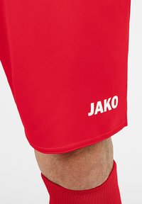 Rote Sportshorts mit glatter Textur, mit einem weißen Logo "JAKO" am unteren Saum. Ein Paar rote Socken ist teilweise sichtbar.