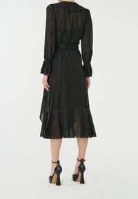 Schwarzes gepunktetes Kleid mit Kragen, langen Ärmeln und einem gerüschten Saum. Kombiniert mit schwarzen High-Heel-Schuhen. Transparenter Stoff verleiht Struktur.