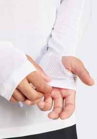 Bijela košulja s dugim rukavima s teksturiranim mrežastim detaljem na manžetama. Mekan materijal s pripijenim rukavima, koji imaju otvore za palčeve radi dodatne zaštite.