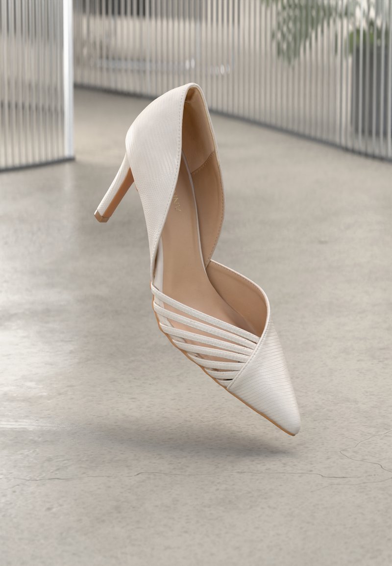 Zapato de tacón alto blanco con punta afilada, textura acanalada y tiras recortadas a los lados, sobre un fondo neutro.