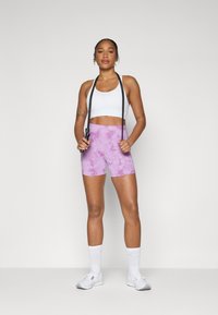Soutien-gorge de sport blanc, shorts tie-dye rose et baskets blanches. Le modèle tient une corde à sauter noire, debout sur un fond uni.