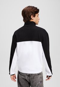 Felpa zip-up bianca e nera con colletto alto, design color-block e maniche lunghe, abbinata a jeans grigi.