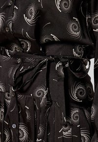 Camicia nera con motivo a pois bianchi a spirale, chiusura con bottoni e cintura annodata in vita. Tessuto morbido con dettaglio gonna plissettata.