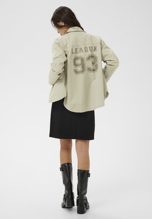 Kobieta w beżowej kurtce z napisem "LEAGUE 93" na plecach, czarnej spódnicy i czarnych wysokich kozaczkach, stojąca na jednolitym tle.