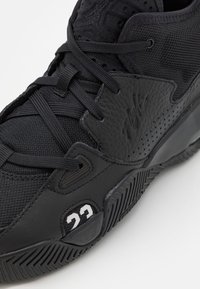 Chaussure de sport noire avec une tige synthétique texturée, un tissu en maille, un système de laçage prononcé et un "23" blanc sur le côté.