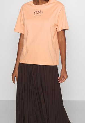 Persoon draagt een perzikkleurig t-shirt met zwarte bloemenborduursels en de tekst "grow wild", gecombineerd met een hoogwaardige geplooide donkerbruine rok met hoge taille.