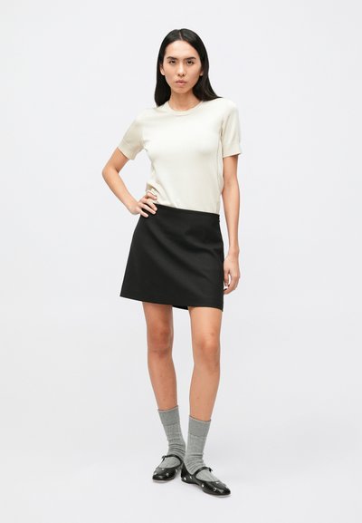 Haut à manches courtes beige clair associé à une jupe trapèze noire. Le mannequin porte des chaussettes grises arrivant aux genoux et des chaussures noires avec des accents décoratifs.