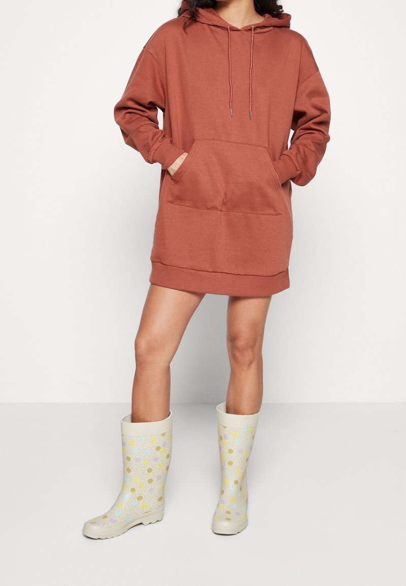 Femme portant une robe sweat à capuche couleur rouille avec les mains dans la poche avant et des bottes de pluie crème à pois multicolores.
