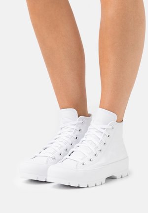 Zapatillas blancas de cuero de caña alta con suelas gruesas y dentadas, usadas en piernas desnudas sobre una superficie blanca.