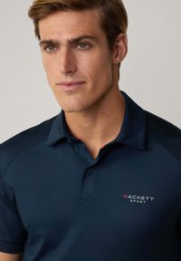 Tummansininen polo paita, jossa on kaulus ja lyhyet hihat. Rinnassa on valkoisena ja punaisena ommeltu logo "HACKETT SPORT".