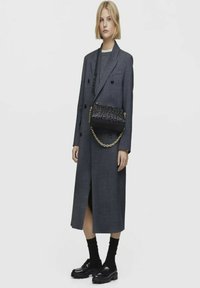 Cappotto lungo grigio con design a doppia abbottonatura, borsa nera con tracolla a catena, calze nere e scarpe nere chunky. Stile minimalista con linee pulite.