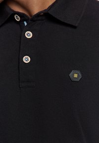 Zwart polo shirt met een gestructureerde stof, een drieknoopskraag met lichtbruine knopen en een zwarte hexagonale patch met een gele accent.