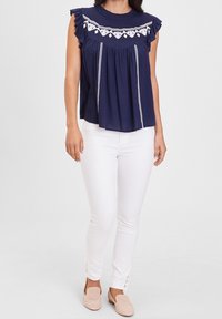 Donna che indossa una canotta senza maniche blu navy con ricami bianchi, pantaloni bianchi attillati con dettagli in bottoni e scarpe slip-on perforate beige.