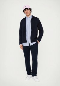 Námořnická bunda na zip přes světle modrou pruhovanou košili, doplněná tmavými kalhotami a bílými teniskami. Růžová čepice na hlavě, jednoduchý design.