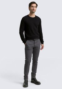 Czarny dziergany sweter z długim rękawem, szare dopasowane spodnie, czarne botki za kostkę, noszone przez mężczyznę stojącego na jasnoszarym tle.