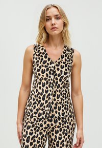 Vrouw in een mouwloze top met luipaardprint en bijpassende broek, staande tegen een effen lichte achtergrond.
