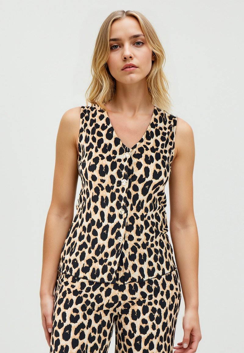 Vrouw in een mouwloze top met luipaardprint en bijpassende broek, staande tegen een effen lichte achtergrond.