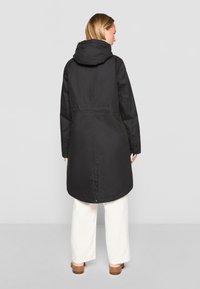 Juodas, su kapišonu, paltas su tiesia forma, ilgomis rankovėmis ir mygtukų detalėmis. Nešiojamas su baltomis plačiomis kelnėmis, lygia tekstūra.