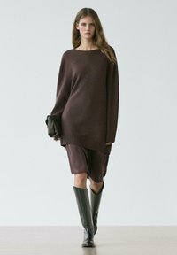 Kobieta idąca w za dużym brązowym swetrze, dopasowanej spódnicy o długości do połowy łydki, czarnych kozakach za kolano, trzymająca czarną kopertówkę na jednolitym tle.