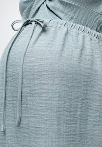 Φόρεμα εγκυμοσύνης ανοιχτού γκρι χρώματος με σούρες στη μέση και δεμένη υφασμάτινη ζώνη, που δείχνει κοντινό στο σημείο της κοιλιάς.