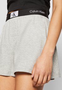 Pantalones cortos de algodón grises con una cinturilla elástica negra que presenta el logo de "Calvin Klein". La tela tiene una textura suave y un ajuste relajado.