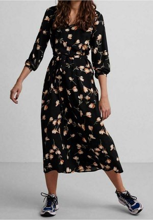 Vrouw draagt een zwarte midi-jurk met bloemenprint, gecombineerd met blauw-witte sportschoenen, staand met gespreide benen tegen een effen achtergrond.