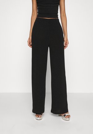 Femme portant un pantalon noir taille haute à jambes larges, un crop top noir ajusté et des sandales à plateforme argentées, debout devant un fond uni.