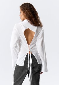 Hvid skjorte med en udskåret ryg, der har krave, lange ærmer og bundet detalje i taljen. Sammen med mørkegrå bukser.