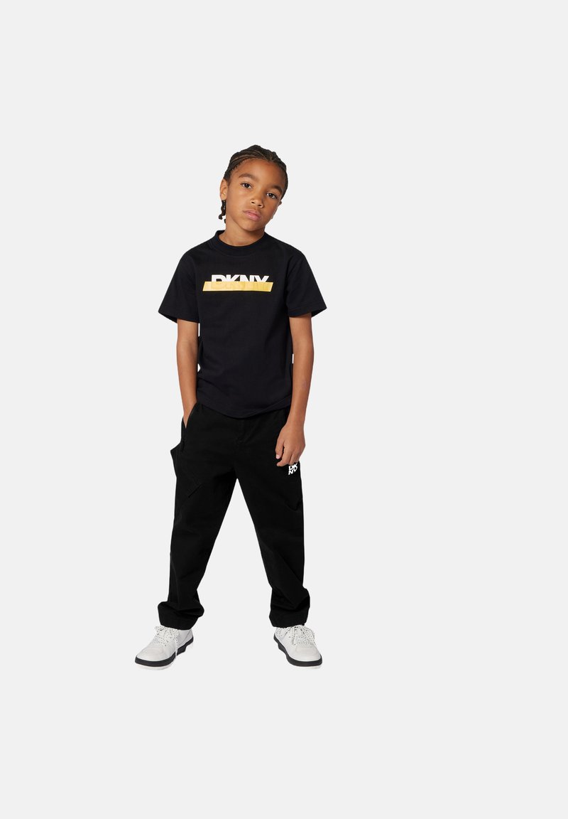 Zwarte t-shirt met een gele DKNY-logo, gecombineerd met zwarte cargo-broek met zakken, en witte sneakers met grijze zolen.