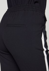Schwarze Sporthose aus strukturiertem Stoff, mit elastischem Bund und einem weißen Seitenstreifen. Mit einem kleinen Rückentaschendetail.