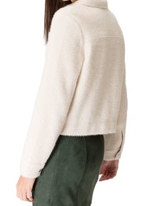 Mujer con una chaqueta de cremoso texturizada, corta y de manga larga, y pantalones verdes de cintura alta, mostrada desde la espalda y el costado.