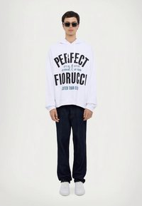 Weißer Hoodie mit großem schwarzen Schriftzug "PERFECT FIORUCCI", kombiniert mit dunkler Jeanshose und weißen Turnschuhen. Das Model steht vor einem hellen Hintergrund.