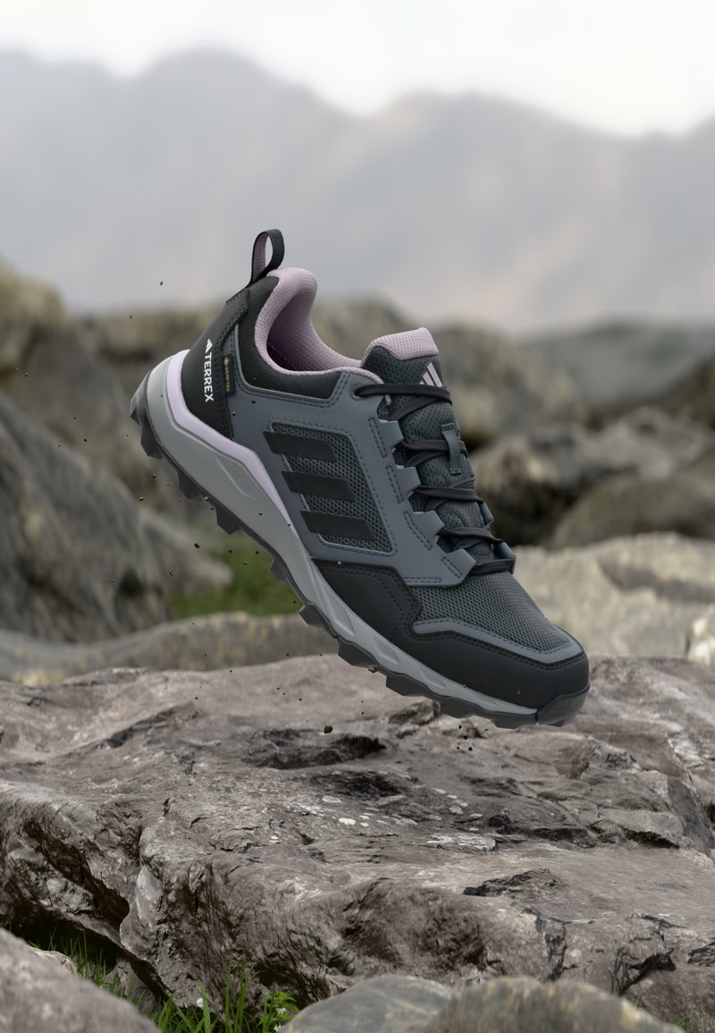 Tummanharmaa ja musta Adidas Terrex -vaelluskengän roikkuu kallioisen maaston yllä, taustalla sumea vuoristomaisema.