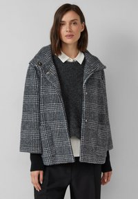 Szary płaszcz w wzór houndstooth z szerokim kołnierzem, dwoma guzikami, bocznymi kieszeniami i luźnym krojem, noszony na ciemnym swetrze i białej koszuli.