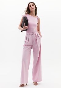 Różowy top bez rękawów z subtelnym falbaną, zestawiony z spodniami o wysokim stanie i szerokich nogawkach oraz czarną torebką. Gładka tekstura, elegancki design.