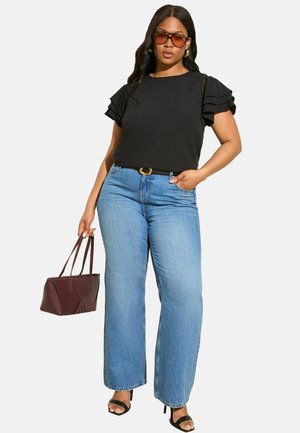 Frau in schwarzem Oberteil mit Rüschensäumen, blauen hochgeschnittenen weiten Jeans, schwarzen Absätzen, brauner Handtasche und braun getönten Sonnenbrille.