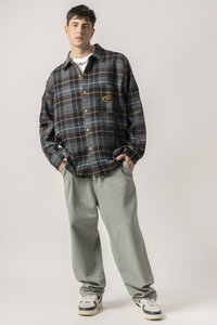 Cămașă în carouri, în nuanțe de navy, gri și ruginie, cu nasturi maronii și o patch cu logo. Asortată cu pantaloni largi de culoare verde deschis și adidași.
