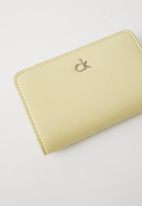 Portefeuille en simili cuir jaune avec bordure cousue et logo "ck" en argent. Forme rectangulaire compacte et texture lisse, design minimaliste.