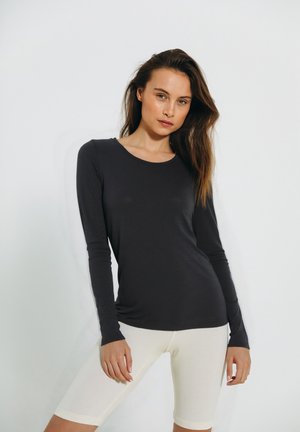 Langærmet, mørkegrå top lavet af blødt stof, parret med lysebeige teksturerede cykel-shorts. Modellen står foran en neutral baggrund.