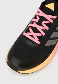 Chaussure de sport en mesh noir avec lacets roses, embout texturé et triangle jaune en accent. Présente des bandes grises sur le côté.