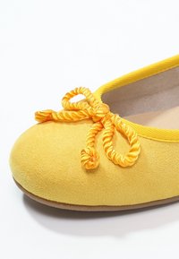Žluté baletní střevíce se semišovou texturou, žlutou šňůrkovou mašlí na špičce a jemným světle hnědým vnitřním podšívkou viditelnou.