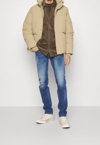Beige puffer jacket over a brown corduroy shirt, paired with blue jeans and cream sneakers. The jacket features a hood and zipper.