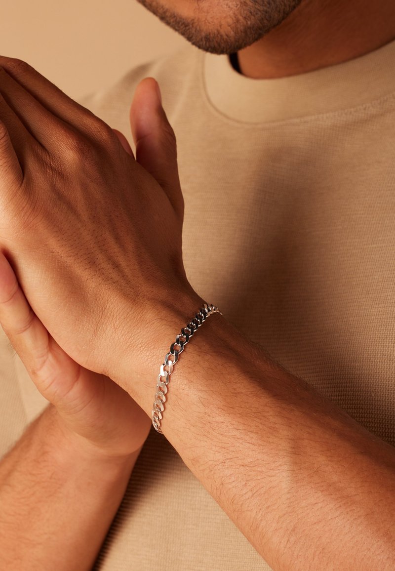 Silver chain bracelet on a wrist, featuring interlocking links with a smooth polish. Hands are positioned together, displaying texture.