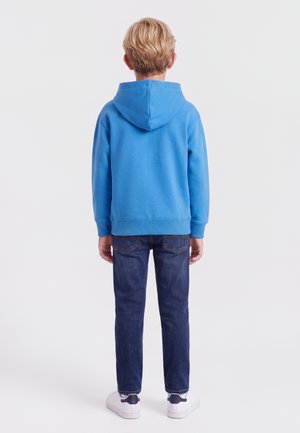 Enfant blond debout de dos, portant un sweat à capuche bleu, un jean bleu foncé et des baskets blanches sur un fond clair uni.