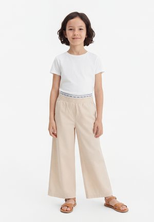 Bambino con capelli corti e scuri, in piedi, indossa una camicia bianca a maniche corte, pantaloni beige a gamba larga e sandali marroni su sfondo bianco.