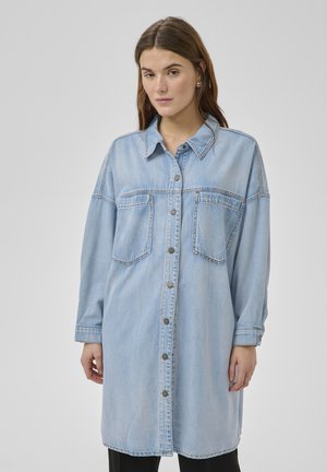 Vrouw draagt een lange lichtblauwe denim blouse met knopen en borstzakken, gecombineerd met een zwarte broek, tegen een effen grijze achtergrond.