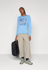 Blue long-sleeve shirt with a mountain graphic, beige pants, gray hiking shoes, and a black backpack, all set against a neutral background.